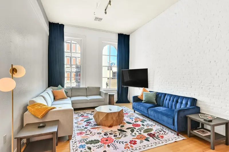 A living room with arched windows, a blue velvet sofa, a gray sectional and a floral rug, sunlight across the floor