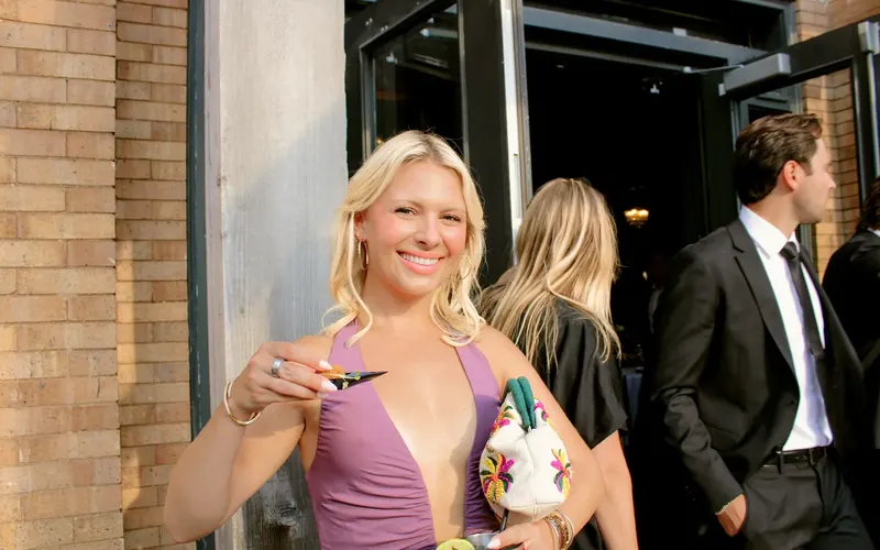 A guest in a purple dress smiling in a doorway, holding a small plate, with other guests behind her