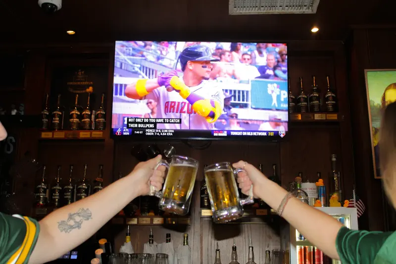 Two beer mugs raised in a toast at the bar, a ball game on the television behind