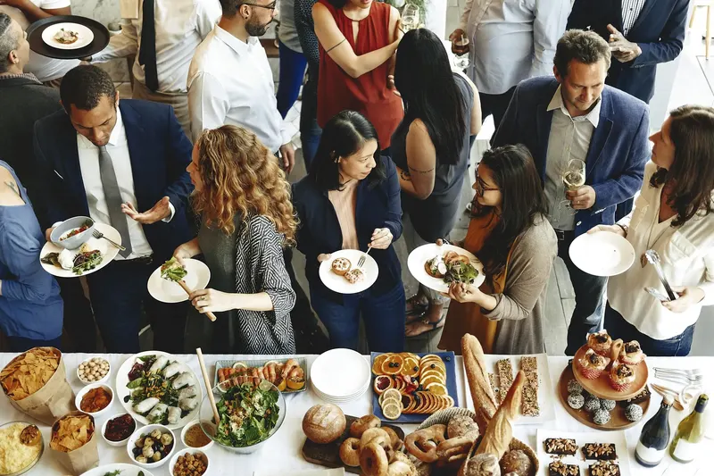 People in blazers and jackets standing with small white plates around a long buffet table of salads, dips, breads and sliced fruit